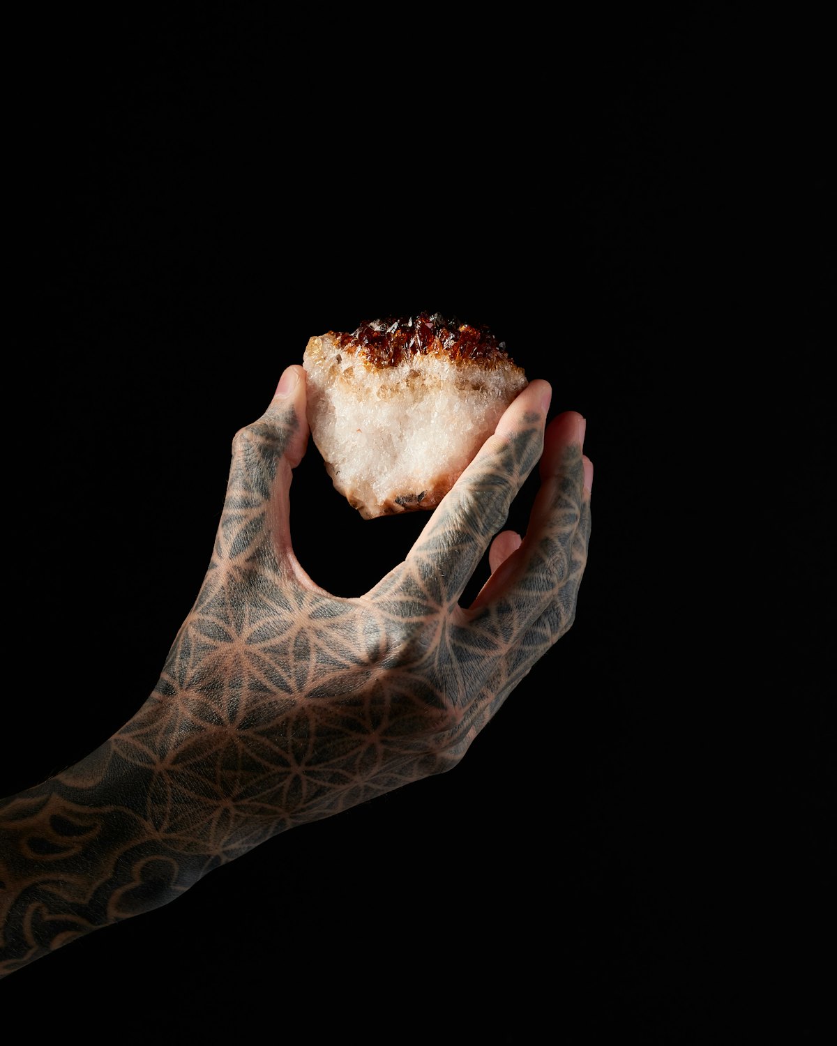 Energy work meets the body where it already speaks — through touch, stone, and attention. A hand tattooed with the Flower of Life, cradling a raw citrine crystal against black