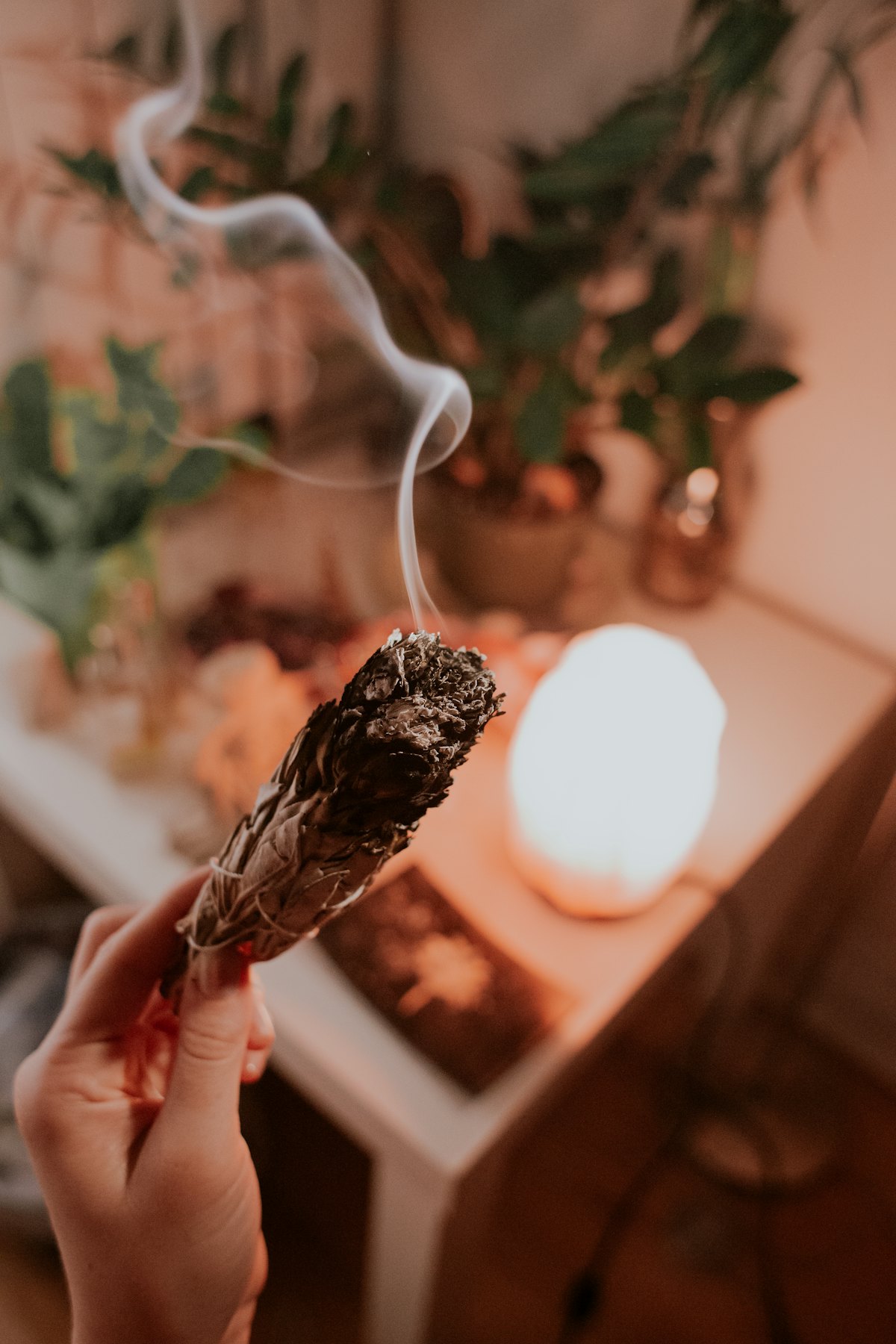 Smudging — clearing the field with smoke and intention before the work begins. A hand holding a smouldering sage bundle, smoke curling up beside a salt lamp